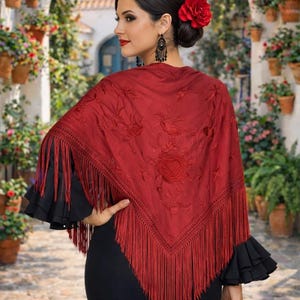 May include: A woman wearing a vibrant red shawl with intricate floral embroidery and long fringe. The shawl is draped over a black dress, and the woman has a red flower in her hair. Black ruffled sleeves peek out from under the shawl.