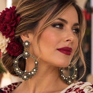 Puede incluir: Un par de pendientes de aro grandes y circulares con acabado en metal dorado antiguo. Cada pendiente presenta un patrón floral de filigrana decorativo acentuado con pequeñas cuentas blancas redondas. Cuelgan de un perno en forma de flor.