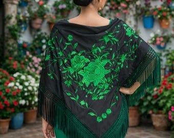 Op de afbeelding: Een zwarte sjaal met groene bloemenborduurwerk en franjes. De sjaal wordt gedragen over een groene jurk. Een groene bloem siert het haar. De achtergrond toont een muur met bloempotten.