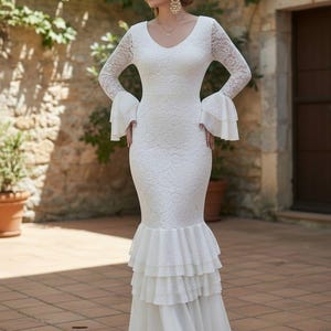 May include: A white lace wedding dress with long sleeves and a V-neck. The dress has a fitted bodice, flared sleeves, and a tiered, ruffled skirt. The dress is floor-length with a slight train.