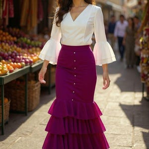 Op de afbeelding: Een vrouw draagt een witte blouse met klokmouwen en een V-hals, gecombineerd met een lange, gelaagde, paarse rok met knoopsluiting. De achtergrond toont een straatbeeld met marktkramen.