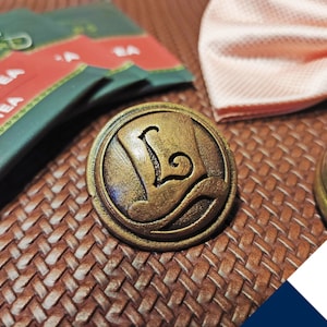 Op de afbeelding: Een gouden metalen pin in de vorm van een hoge hoed met de letter "L" aan de zijkant. De pin is op een bruin geweven oppervlak.