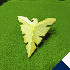 May include: A gold-colored metal eagle pendant with a geometric design. The pendant is on a green surface.