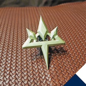 Peut inclure: Une épingle en plastique imprimée en 3D de couleur verte en forme d'étoile avec un point en haut et quatre points en bas. L'étoile a un design géométrique avec un point central et quatre points plus petits autour.