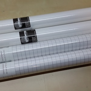 Könnte beinhalten: Vier Rollen weißes Geschenkpapier, zwei unifarben und zwei mit einem Rastermuster. Die Rollen sind zylindrisch und in Klarsichtfolie verpackt. Das Rasterpapier hat ein schwarz-weißes Design. Die Rollen liegen auf einer hellbraunen Oberfläche.