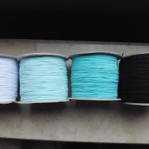 Pode incluir: Quatro rolos de cordão elástico em branco, azul claro, turquesa e preto. Cada rolo é enrolado firmemente e assenta sobre um núcleo de cartão. Os cordões são provavelmente usados para projetos de costura ou artesanato.