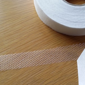 May include: A roll of white interfacing tape with a clear, open-weave adhesive strip. The tape is partially unrolled, showing the adhesive. The background is a light-coloured wooden surface.