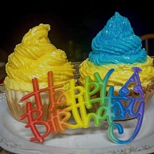 May include: Two cupcakes with yellow and blue frosting, presented on a white plate. A rainbow-colored "Happy Birthday" decoration is in front of the cupcakes. The cupcakes are in brown paper liners.