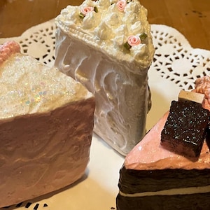 May include: Three decorative cake slices. One slice is pink with a glittery top and a pink border. Another is white with white frosting and pink roses. The third slice is pink and brown with chocolate squares.