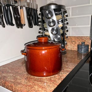 May include: A glazed, reddish-brown ceramic cooking pot with a lid and side handles. The pot sits on a speckled countertop near a spice rack and a stovetop. Kitchen utensils hang on the wall in the background.