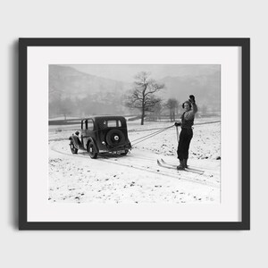 May include: Black and white framed print of a vintage car pulling a skier across a snowy field. The skier is wearing dark clothing and skis, waving their arms. The car has a spare tire on the back.