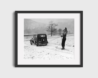 May include: Black and white framed print of a vintage car pulling a skier across a snowy field. The skier is wearing dark clothing and skis, waving their arms. The car has a spare tire on the back.