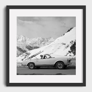 Könnte beinhalten: Ein Schwarzweißfoto eines Oldtimers, der durch eine schneebedeckte Berglandschaft fährt. Das Auto hat Skier auf dem Dach befestigt und zwei Personen sitzen darin.