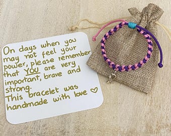 May include: A handmade bracelet with pink and purple braided cord, a small charm, and a drawstring bag. The bracelet is next to a card with the text: "On days when you may not feel your power, please remember that YOU are very important, brave and strong."
