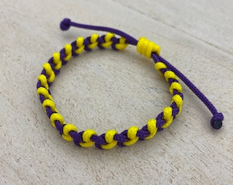 Könnte beinhalten: Ein geflochtenes Freundschaftsarmband in Lila und Gelb. Das Armband besteht aus zwei Schnüren, einer lila und einer gelben, die miteinander verflochten sind, um ein Muster zu erzeugen.