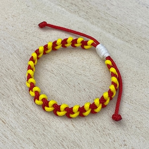 May include: A red and yellow braided friendship bracelet with a white knot. The bracelet is made of two strands of cord that are woven together.