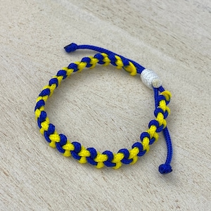 May include: A yellow and blue braided friendship bracelet with a white bead. The bracelet is made of two strands of cord that are woven together.
