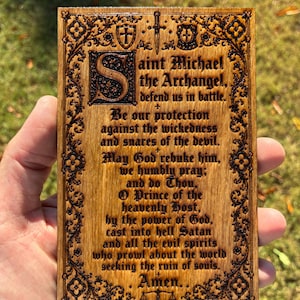 May include: A rectangular wooden plaque featuring a laser-engraved prayer to Saint Michael the Archangel. The text is surrounded by an ornate floral border with a sword, shields, and scales. The wood grain is visible, and the engraving is dark brown.
