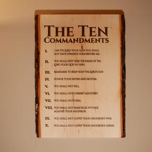May include: A wooden plaque with the text "THE TEN COMMANDMENTS" in a dark brown font. The text is centered at the top, with the commandments listed below in a similar font. The wood has a natural edge.