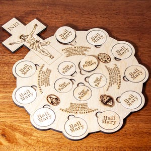 May include: A wooden rosary board with laser-cut text and images. The board features circular cutouts with the words "Hail Mary" and "Our Father." A wooden cross with the letters "INRI" and an image of Jesus is included.