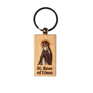 May include: A wooden keychain featuring an image of St. Rose of Lima. The rectangular pendant has a black ring and a silver-colored metal connector. The image depicts St. Rose in a black cloak and white veil, with the text "St. Rose of Lima" below.