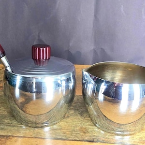 May include: A vintage silver-toned sugar bowl with a lid and a matching creamer. The sugar bowl has a red-handled spoon, and the creamer has a red handle. Both pieces are reflective and sit on a wooden surface.