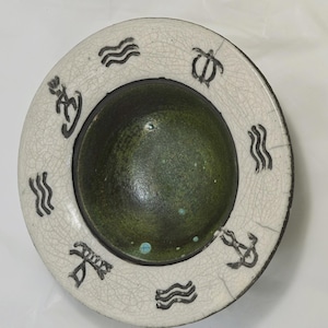 Peut inclure: Un bol en céramique avec un intérieur vert foncé et un bord blanc. Le bord présente des symboles nautiques noirs et une texture craquelée. Le design du bol comprend des vagues, des ancres et une tortue.