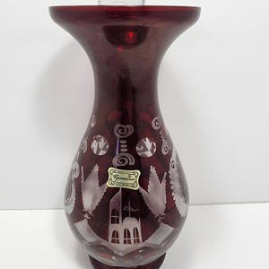 May include: A ruby red glass vase with etched white floral and architectural designs. The vase has a flared top and a rounded base. A small label is affixed to the front. The vase is approximately 20 cm tall.