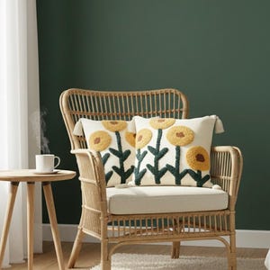 May include: A natural rattan armchair with a cream-colored cushion and two decorative pillows. The pillows feature a yellow flower design with green stems on a cream background. A small wooden side table with a white mug sits nearby.