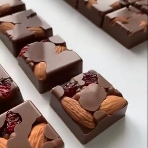 Könnte beinhalten: Eine Reihe von quadratischen Zartbitterschokoladenstücken, die in Reihen angeordnet sind. Jedes Stück hat eine glatte Schokoladenbasis, die mit ganzen braunen Mandeln und getrockneten roten Cranberries belegt und teilweise mit geschmolzener Schokolade überzogen ist.