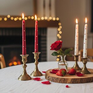 Könnte beinhalten: Eine romantische Tischdekoration mit brennenden Kerzen, einer Rose und Erdbeeren. Rote Kerzen mit weißen Herzmustern in Messingkerzenhaltern. Eine rote Rose und Erdbeeren liegen auf einer Holzscheibe, mit verstreuten Rosenblättern.