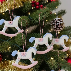 Puede incluir: Adornos navideños de caballo balancín blancos, con crines azules y balancines rosas. Los adornos están decorados con detalles rosas y azules y cuelgan de un cordel rojo y blanco en un árbol de Navidad decorado con bolas rojas y doradas.