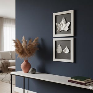 Peut inclure: Deux œuvres d'art encadrées avec des motifs de feuilles argentées. Les feuilles sont placées sur un fond gris foncé dans des cadres blancs. Un vase avec de l'herbe de la pampa séchée et une table décorative avec des livres et une petite sphère complètent le décor.