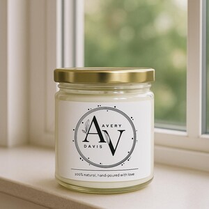 Op de afbeelding: Een glazen potkaars met een gouden deksel. Het etiket toont de tekst "AVERY DAVIS" in een cirkelvormig ontwerp met botanische accenten. De onderkant van het etiket luidt "100% natuurlijk, met de hand gegoten met liefde."