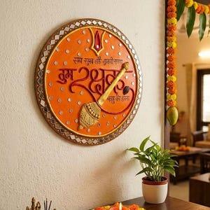 Puede incluir: Una placa de pared circular naranja con una maza dorada en relieve, un símbolo tilak rojo y el texto en hindi "सब सुख लहै तुम्हारी सरना, तुम रक्षक काहू को डरना". El borde está decorado con pequeños espejos cuadrados y un adorno dorado.