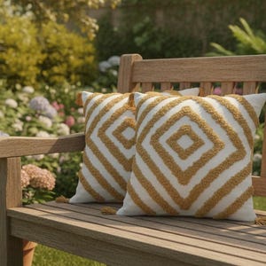 Op de afbeelding: Twee decoratieve sierkussens met een geometrisch ruitpatroon in goud en wit, liggend op een houten bank. De kussens hebben een textuurontwerp en kleine kwastjes. De bank staat buiten in een tuin.