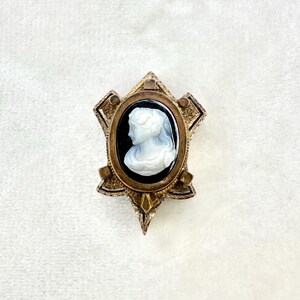 May include: Vintage cameo brooch featuring a white profile against a black background, set in an oval frame. The frame is surrounded by an ornate, gold-toned metal design with a starburst shape. A classic piece of jewelry.