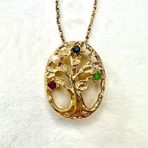 May include: Gold-toned pendant necklace featuring an oval-shaped tree design. The tree has colorful gemstones: a pearl, a ruby, a sapphire, and an emerald. The necklace is on a gold chain.
