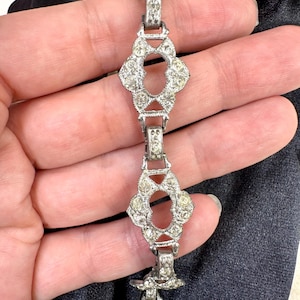 May include: Silver-toned bracelet with a series of ornate, diamond-shaped links. Each link is embellished with small, clear gemstones. The bracelet is shown against a dark background, highlighting its intricate design and sparkle.