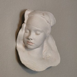 May include: White plaster sculpture of a woman's face with closed eyes. The sculpture is mounted on a wall.