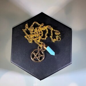 May include: A gold chain necklace with a pentagram pendant and a light blue crystal charm. The necklace is displayed on a black velvet background.