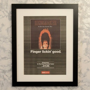 Può includere: Pubblicità incorniciata per Kingmaker, con cornice nera e passe-partout bianco. L'immagine presenta una persona con le braccia alzate e il testo "Finger lickin' good." Lo sfondo è marmorizzato.