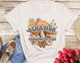 May include: A white t-shirt with a graphic design of mushrooms and flowers. The text "BEHAVIOR is COMMUNICATION" is printed on the shirt.