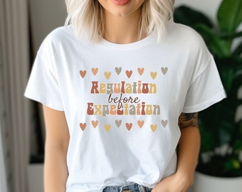 May include: White t-shirt with the text "Regulation before Expectation" in a retro font with brown, orange, and beige colors. The text is surrounded by a row of small hearts in various shades of brown, orange, and beige.