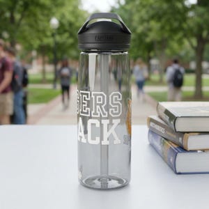 May include: A clear water bottle with a black lid and handle, featuring the text "Tigers Back" in white block letters. The bottle has a straw and a logo on the lid. The background shows a blurred outdoor scene with people and trees.