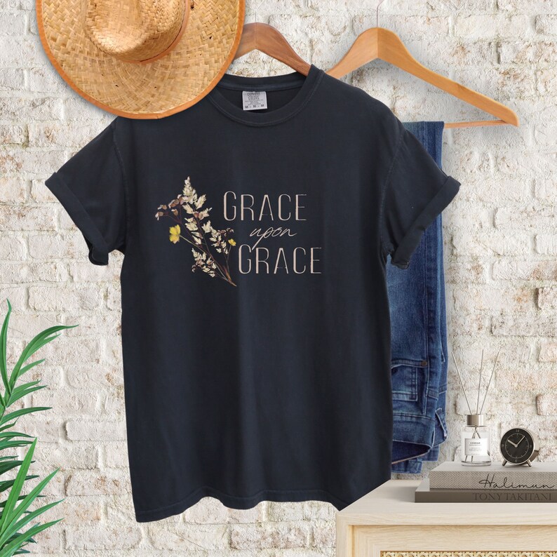 Peut inclure: Un t-shirt noir avec un motif floral et le texte "GRACE upon GRACE" en brun clair.