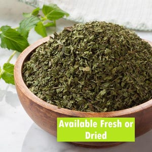 May include: A wooden bowl filled with dried green herbs, possibly mint, is on a white surface. Fresh mint sprigs are to the left. Text on a green rectangle reads "Available Fresh or Dried".