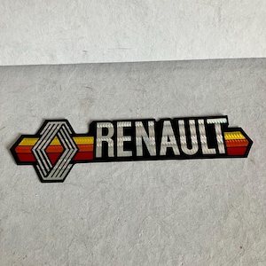 Könnte beinhalten: Ein schwarzes und silbernes Renault-Emblem mit geometrischem Design und dem Markennamen in Großbuchstaben. Das Emblem hat einen Farbverlauf aus Rot, Orange und Gelb.