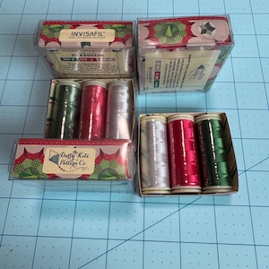 May include: A collection of sewing thread spools in clear plastic boxes. The spools are in shades of green, red, and silver. The boxes have decorative labels with text that reads "INVISAFIL" and "Darty Kite Pattern Co."