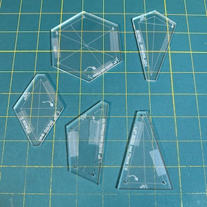 May include: A set of clear acrylic quilting templates in diverse geometric forms, such as a hexagon, diamond, and triangles. Each template has etched markings and measurements in inches, with a grid pattern for accurate cutting. The templates are displayed on a green cutting mat.